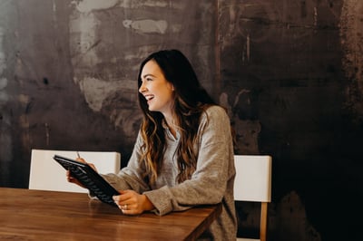 A white woman is sitting at a large, dark wood table. She's holding a tablet computer, looking off towards a window on her left. She has long brown hair, and is laughing.