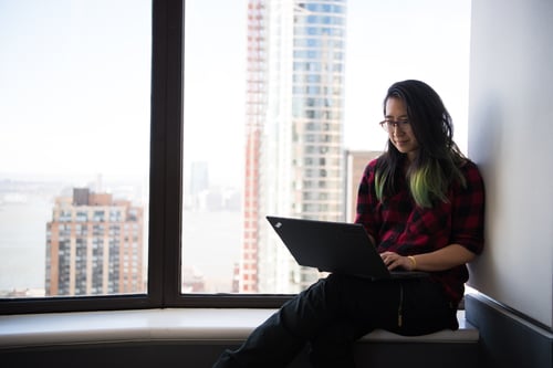 White woman leaning on a wall next to a large window. She is concentrating on her laptop and working.