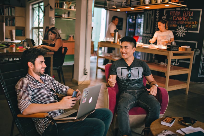 Two young men, one south Asian and one white, sit in a casual, artfully cluttered work space, smiling and talking about work.